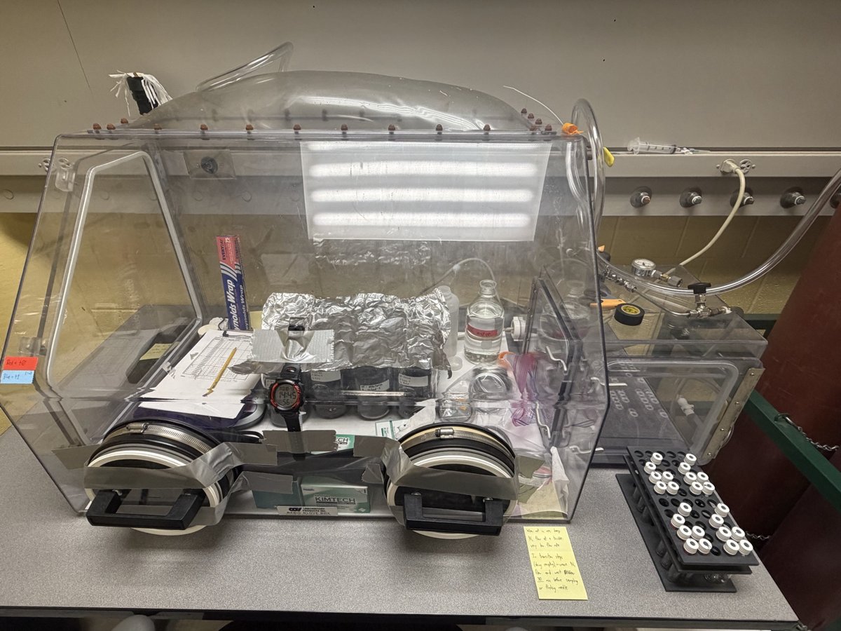 Clear acrylic laboratory glovebox with glove ports and sealed jars inside, on a lab bench.