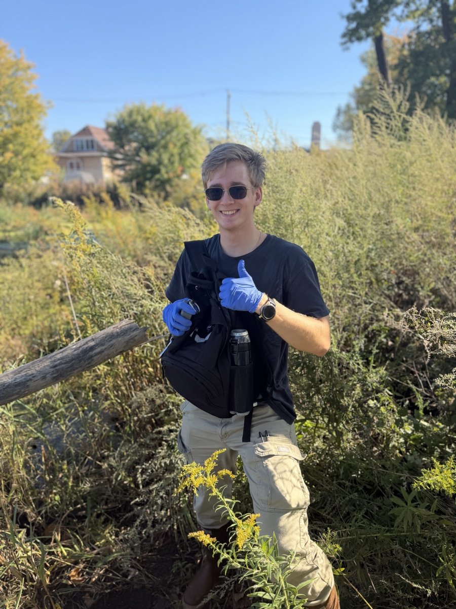 Smiling student in sunglasses and blue nitrile gloves giving a thumbs-up while standing in tall goldenrod and grasses at a marsh, carrying a field bag.